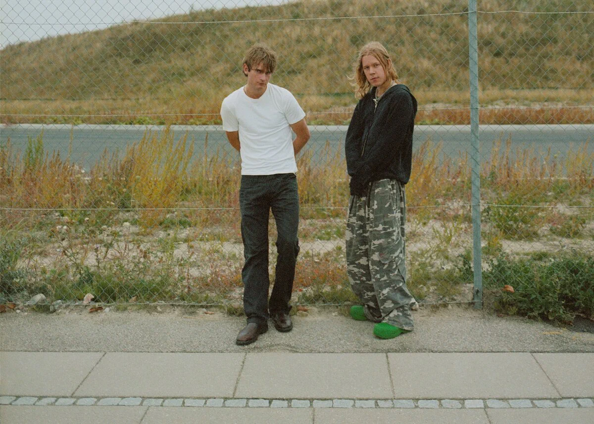 A Good Year, two men standing side by side in front of a wire fence and a grassy embankment, one in a white T-shirt and one in a dark jacket and camouflage trousers