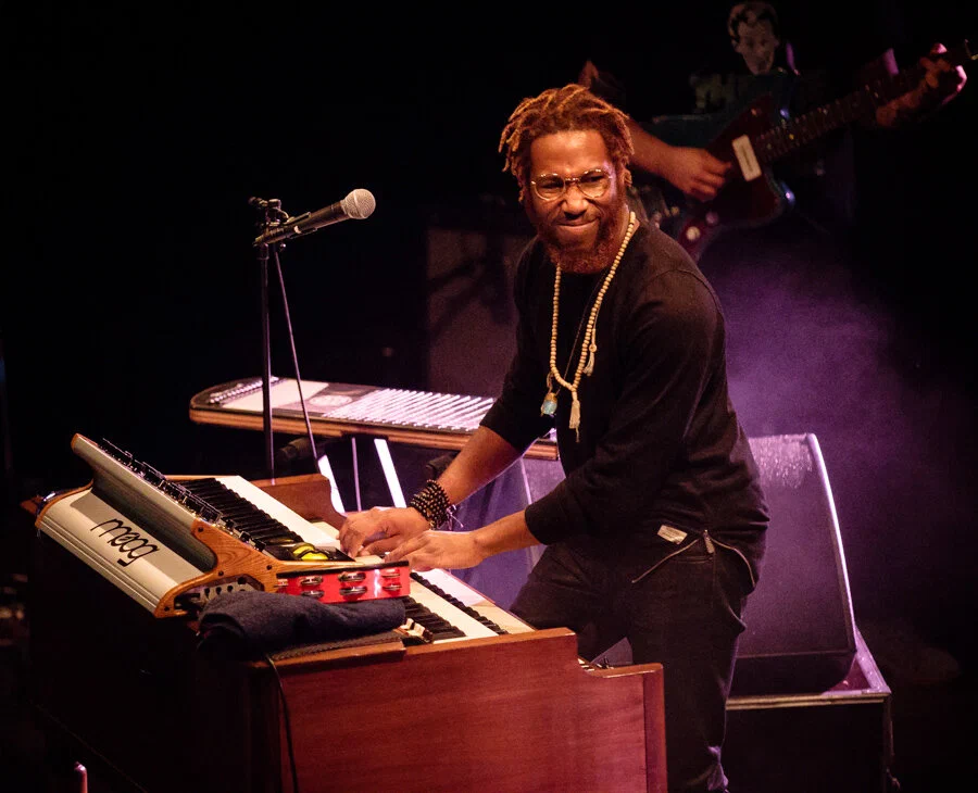 Cory Henry smiling at an organ with a Moog keyboard on top, on a dark stage