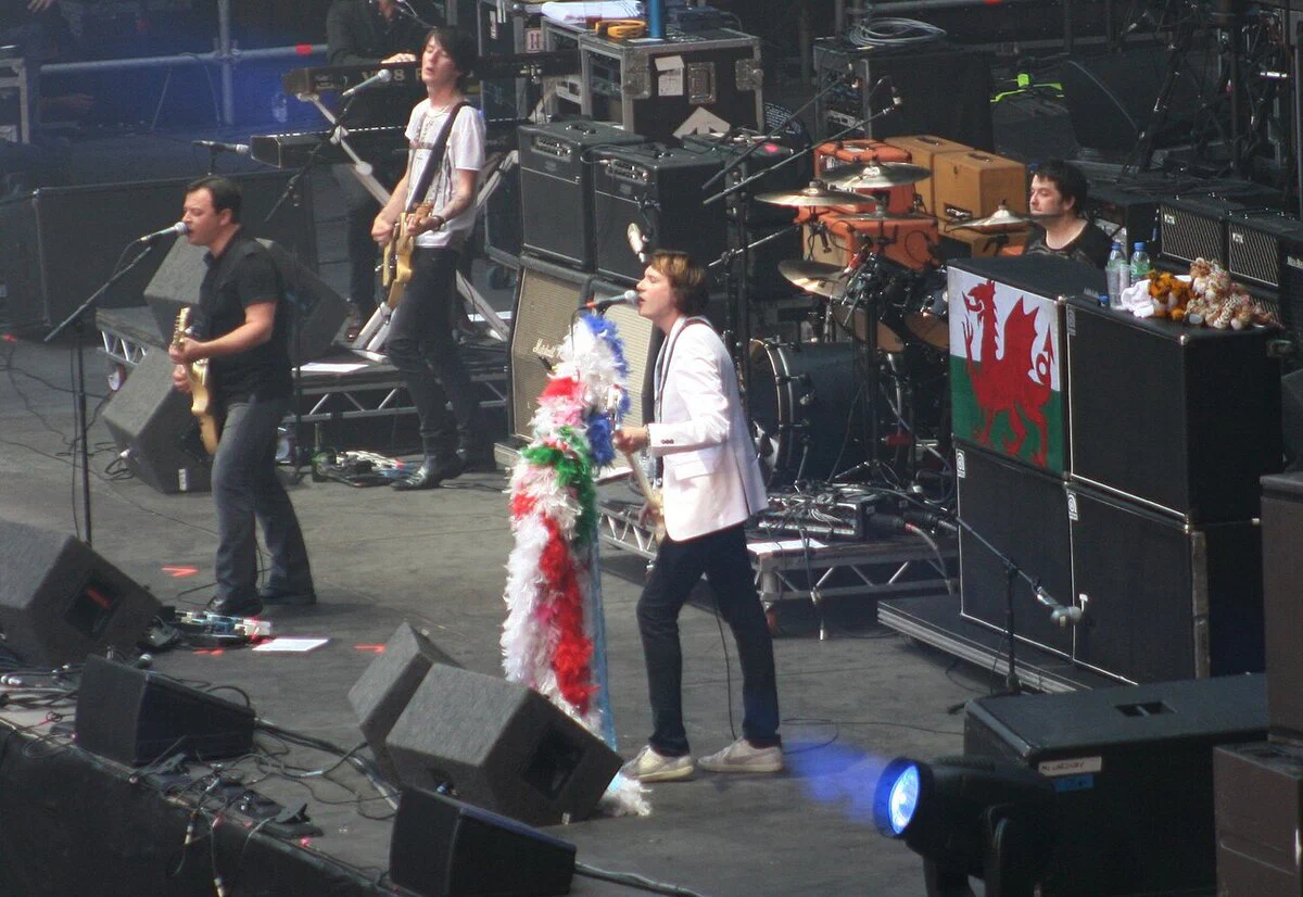 Manic Street Preachers performing on a large stage in Cardiff, with a singer-guitarist, a singer in a white jacket, a guitarist and a drummer behind a Welsh flag on an amplifier