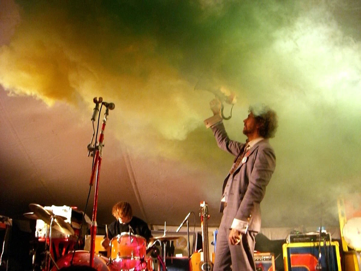The Flaming Lips on stage, with singer Wayne Coyne holding a megaphone aloft beside the drum kit under green and yellow stage smoke