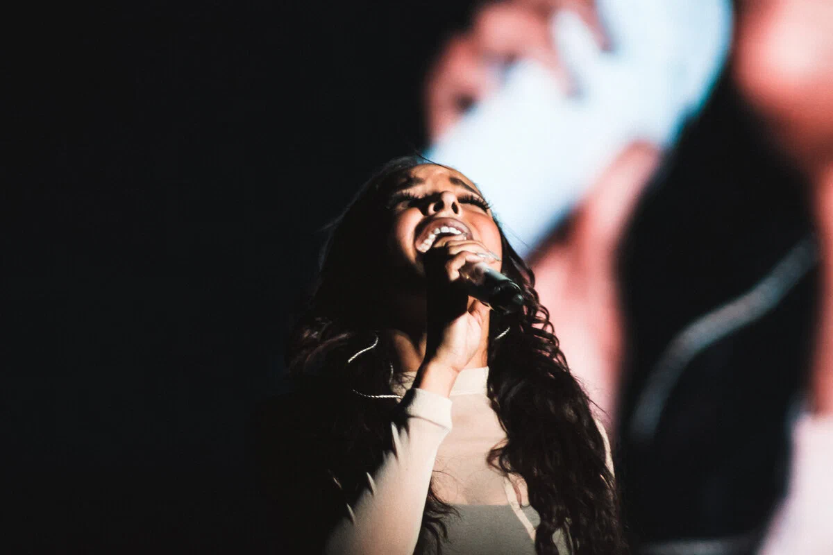Tinashe singing into a microphone with her head tilted back during a concert, in front of a dark video screen