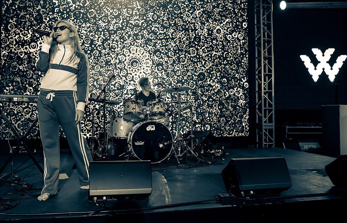 Ashe singing into a microphone on stage beside a drummer in front of a patterned video screen