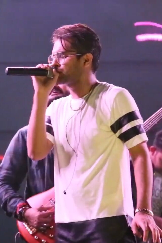 Singer in glasses and a white T-shirt performing into a microphone on a dimly lit stage with a guitarist behind him