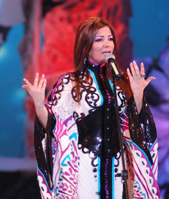 Singer Assala Nasri performing on stage at a microphone in a patterned embroidered gown