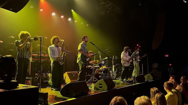 Six-piece band with trumpet, saxophone, guitars and drums playing under yellow and orange stage lights