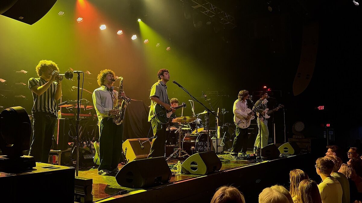 Six-piece band with trumpet, saxophone, guitars and drums playing under yellow and orange stage lights