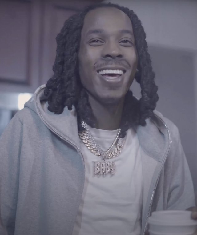 Smiling man with locs in a gray zip hoodie and a diamond chain pendant reading BABY