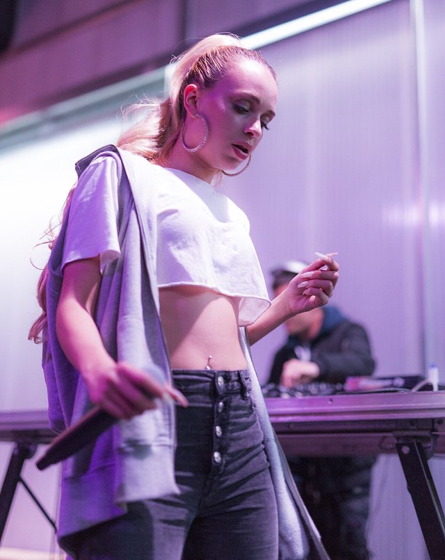 Bad Gyal performing on stage in a white crop top with a DJ behind her under purple light