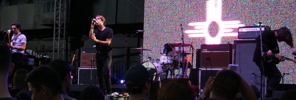 Bad Suns performing on an outdoor stage, singer at the microphone with a guitarist and drummer behind a crowd