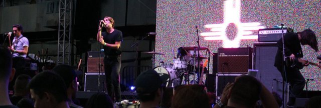 Bad Suns performing on an outdoor stage, singer at the microphone with a guitarist and drummer behind a crowd