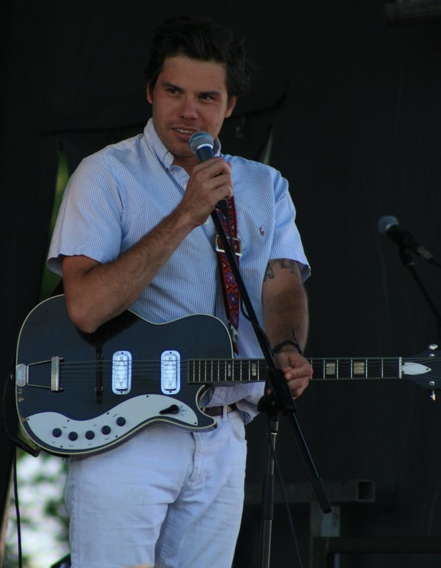 Singer holding a microphone and wearing a black electric guitar on an outdoor stage
