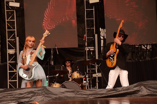 Three members of a band on an outdoor festival stage, two holding guitars and one seated behind a drum kit