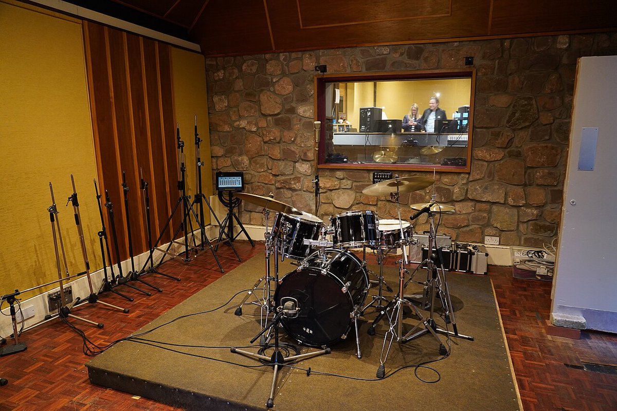 An empty recording studio live room with a drum kit set up in front of a stone wall and a window into the control room