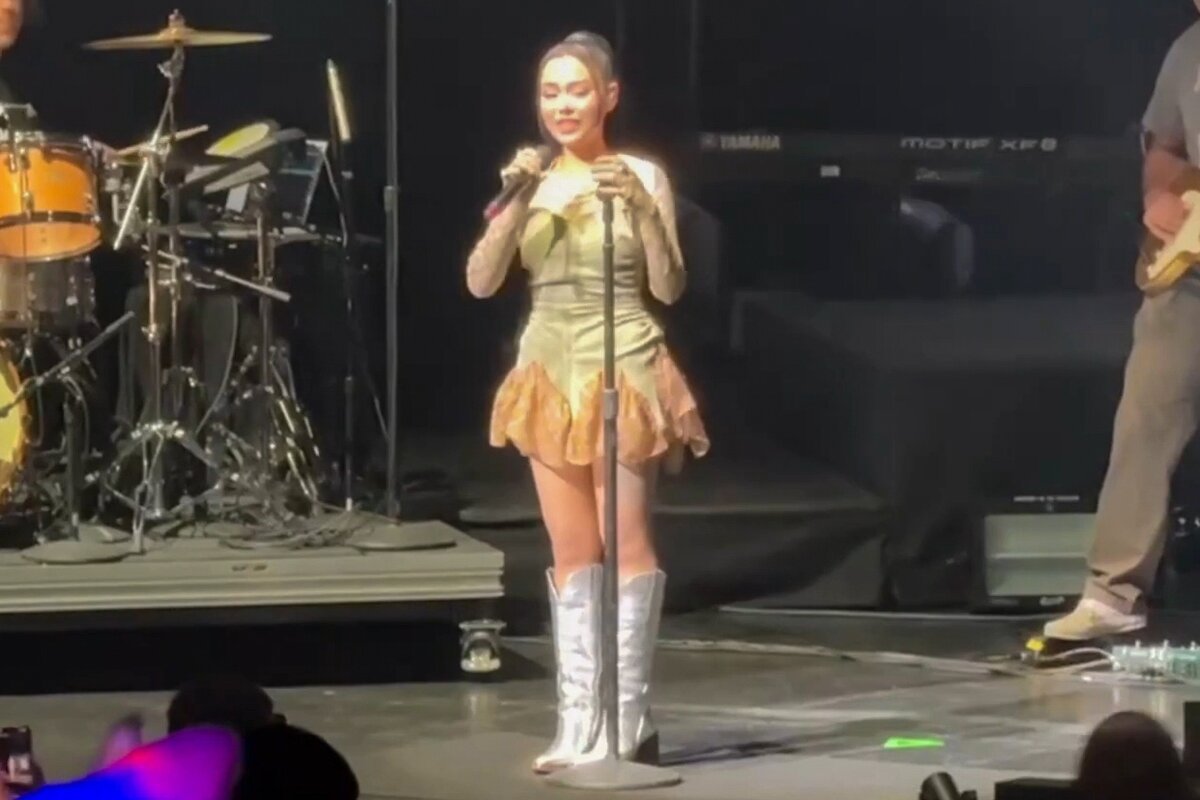 Bella Poarch singing into a microphone on stage in a sparkly mini dress and silver boots, with a drum kit behind her