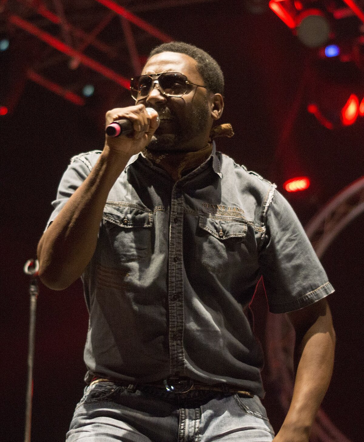 Big Daddy Kane in tinted aviator glasses rapping into a microphone on a stage lit with red lights