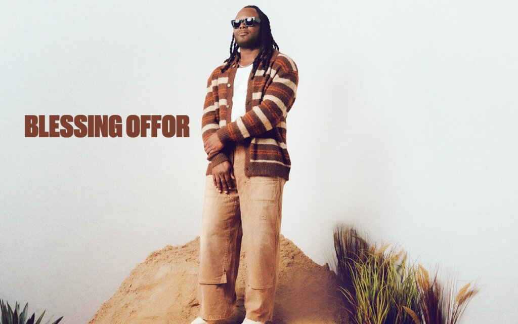 Blessing Offor standing in sunglasses and a striped cardigan against a pale backdrop with his name in brown lettering