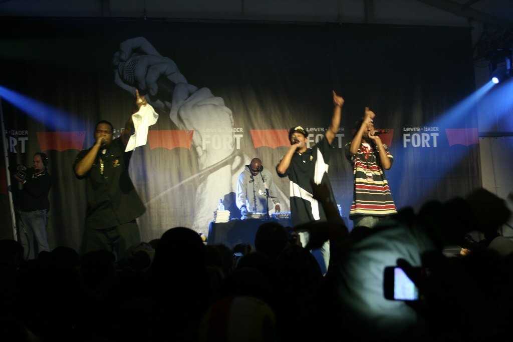 Three members of a hip-hop group rapping on a dark stage under blue lights, with a silhouetted crowd in the foreground