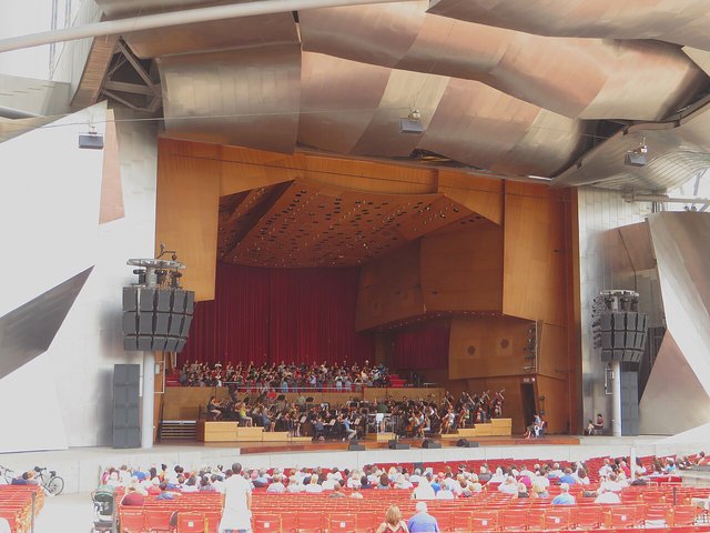 The curved wooden stage shell of an outdoor concert pavilion with an orchestra seated inside, seen from the lawn