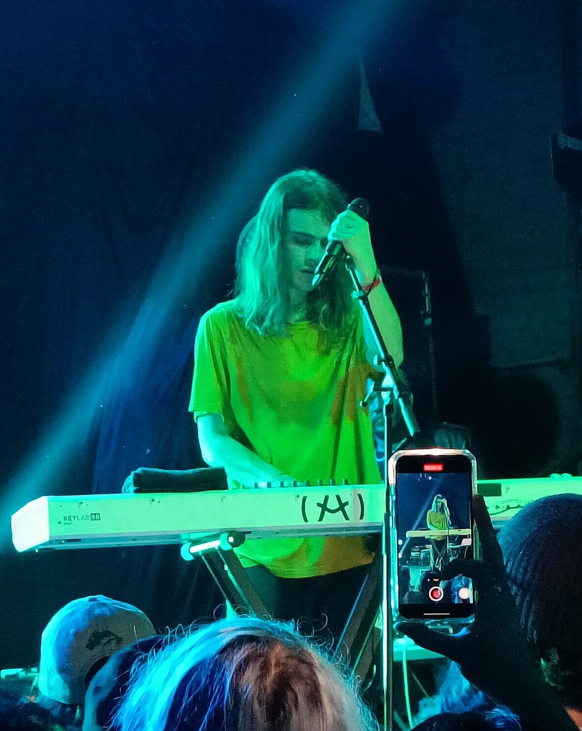 Brakence singing into a microphone behind a keyboard under green stage light, with a fan filming on a phone in the foreground