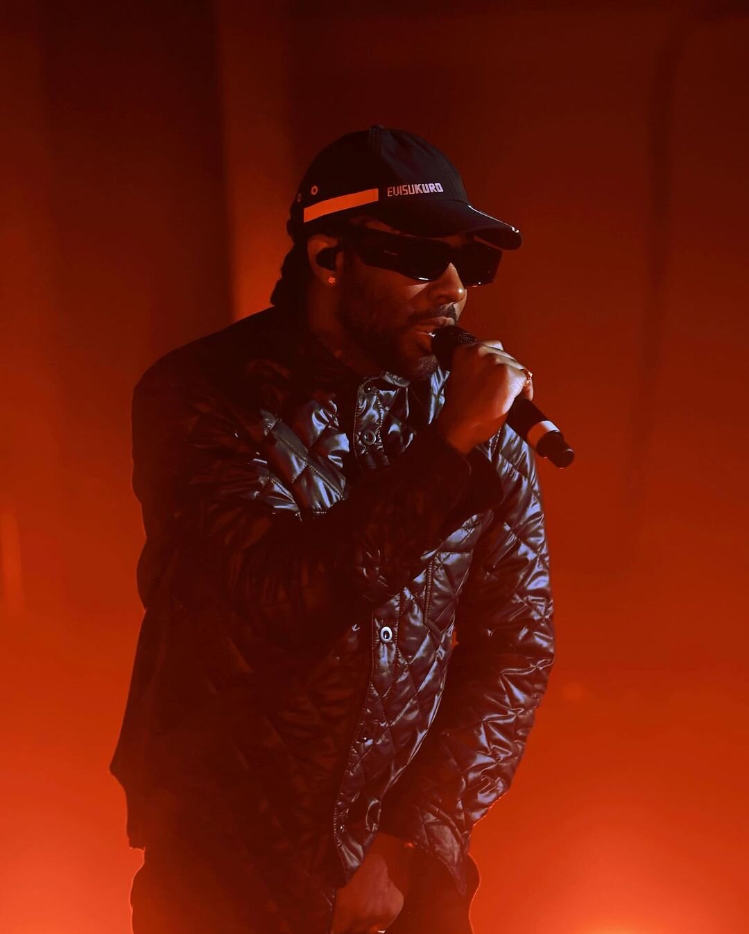 Brent Faiyaz singing into a microphone in a black cap, sunglasses and quilted jacket, lit in orange stage haze