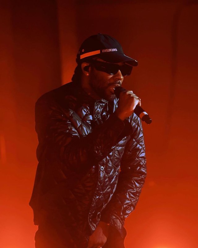 Brent Faiyaz singing into a microphone in a black cap, sunglasses and quilted jacket, lit in orange stage haze