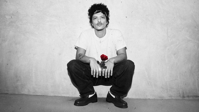 Bruno Mars in a bandana and white T-shirt crouching against a gray wall, holding a single red rose