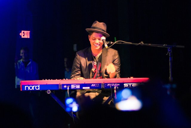 Bruno Mars in a fedora smiling while playing a red Nord keyboard on a dark stage