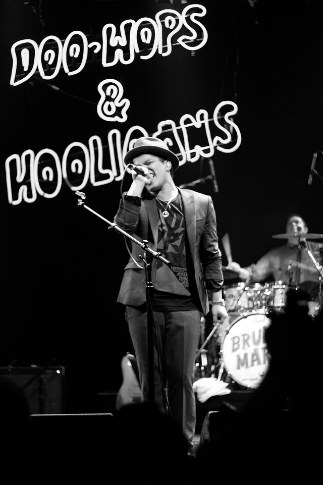 Bruno Mars singing into a microphone on stage in front of a Doo-Wops and Hooligans neon sign