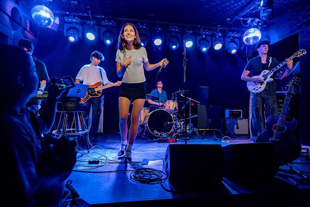 A band on a small club stage lit in blue, with the audience's heads in the foreground