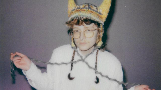 Cavetown in round glasses and a hand-knit horned hat, holding a strand of gray yarn against a lavender wall