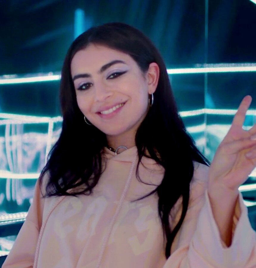 Smiling woman with long dark hair and winged eyeliner in a peach hoodie, making a peace sign in front of teal neon lights