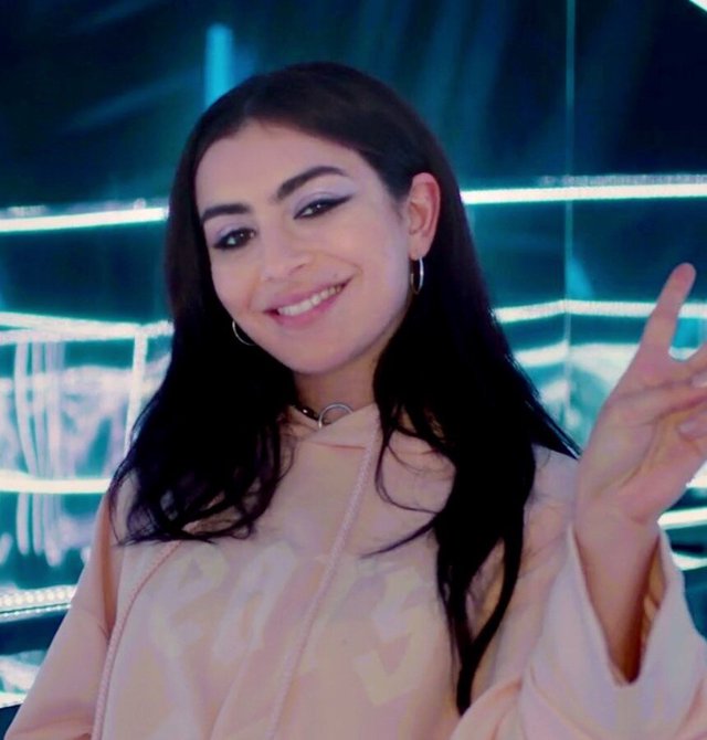 Smiling woman with long dark hair and winged eyeliner in a peach hoodie, making a peace sign in front of teal neon lights