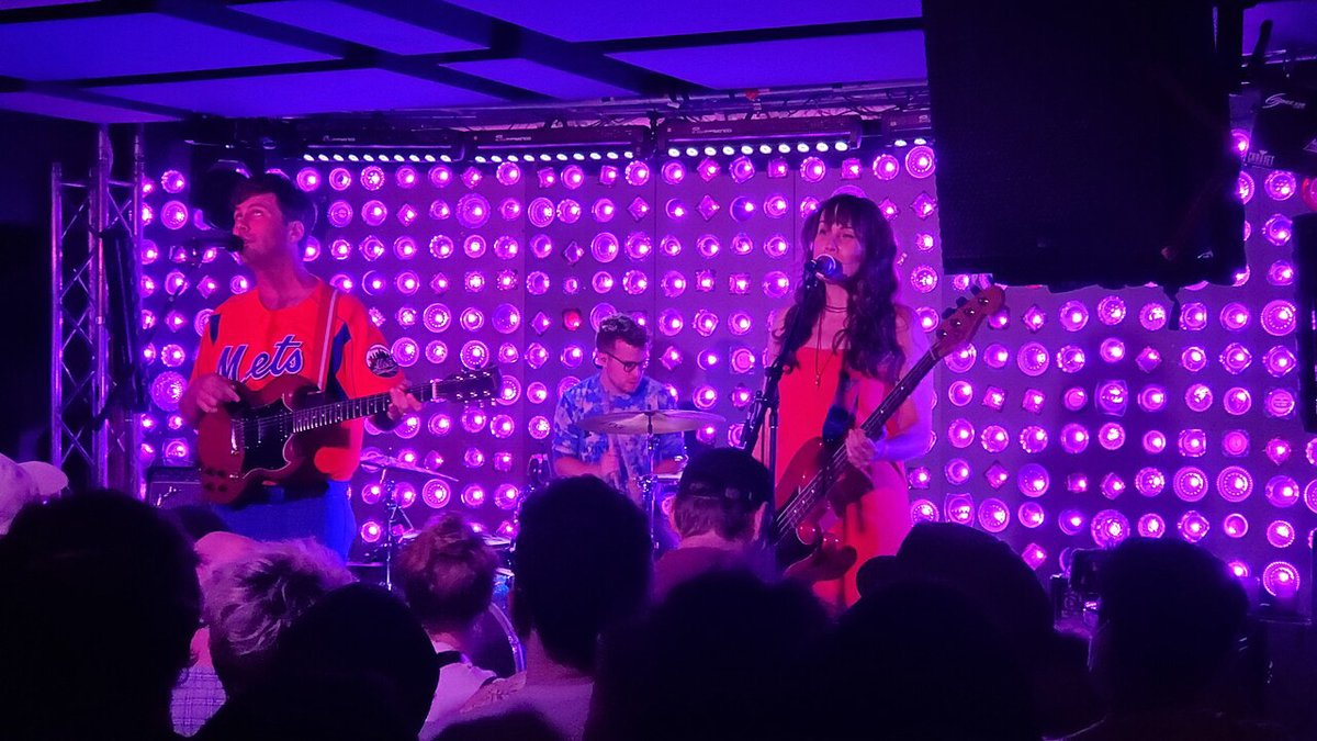 Three-piece band playing on a stage lit by a wall of purple bulbs, with the audience in silhouette