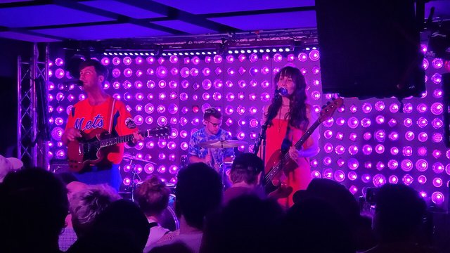 Three-piece band playing on a stage lit by a wall of purple bulbs, with the audience in silhouette