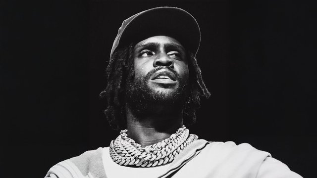 Black and white photo of Chief Keef in a ball cap and stacked chain necklaces, looking up from the stage