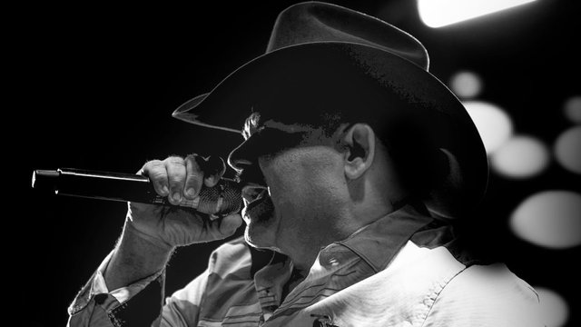 Black-and-white photo of Chuy Lizarraga in a cowboy hat singing into a microphone under stage lights