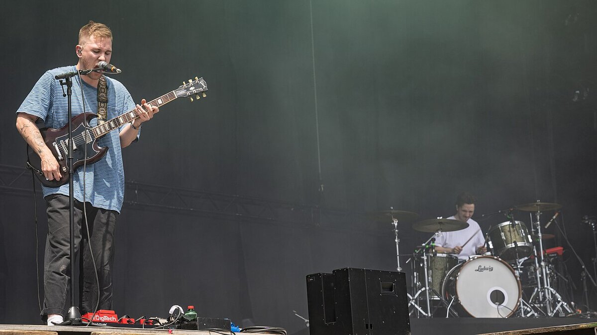 Guitarist singing at a microphone on a festival stage with a drummer behind him