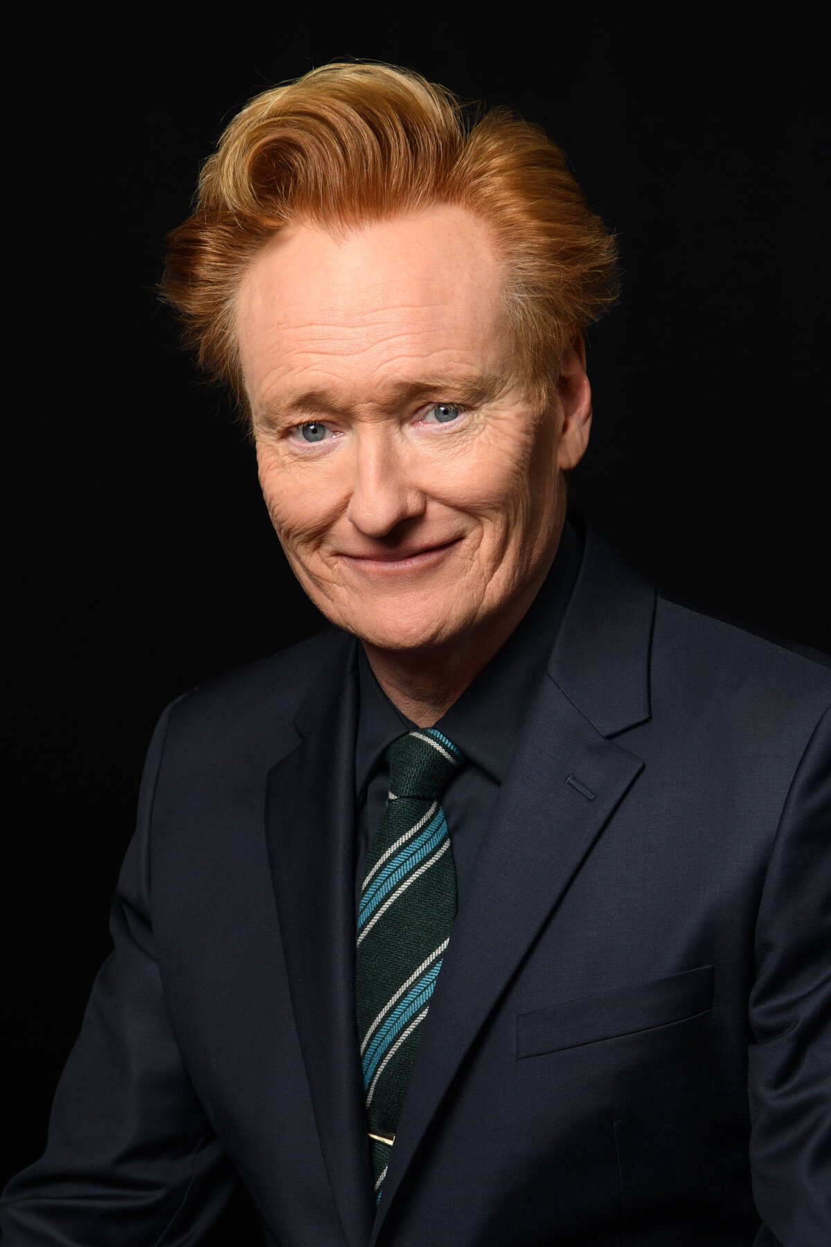 Smiling Conan O'Brien with swept-up hair, wearing a dark denim jacket and a lanyard