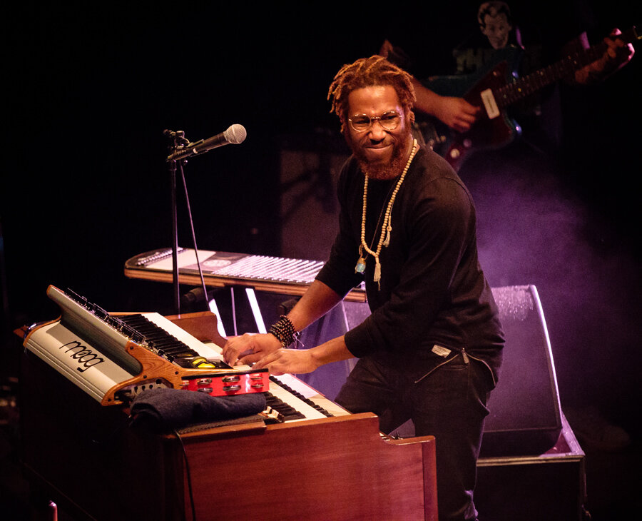 Cory Henry smiling at a Moog keyboard and organ on a dimly lit stage