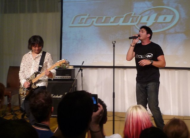 Two members of Crush 40 performing on a small stage in front of a screen showing the band logo