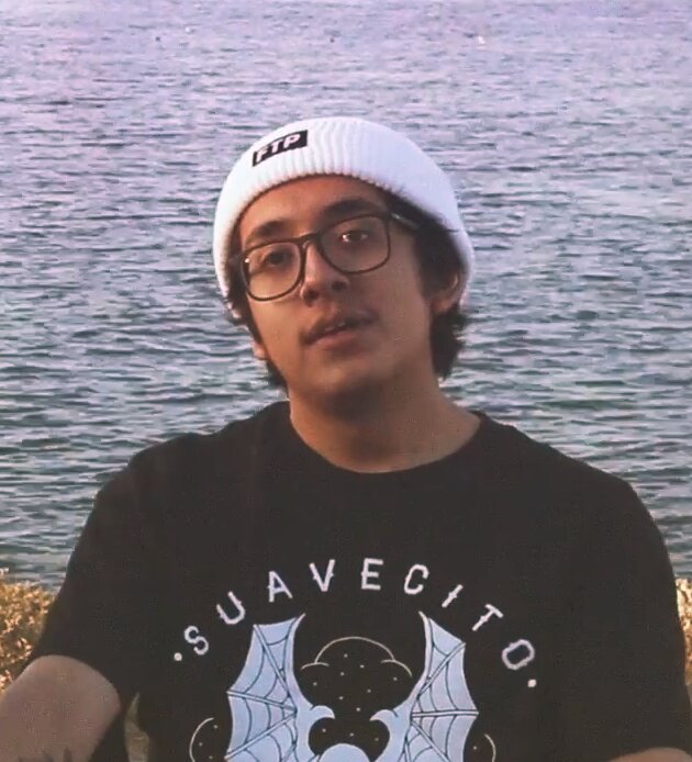 Cuco in a white beanie and glasses, wearing a black graphic tee, with water behind him