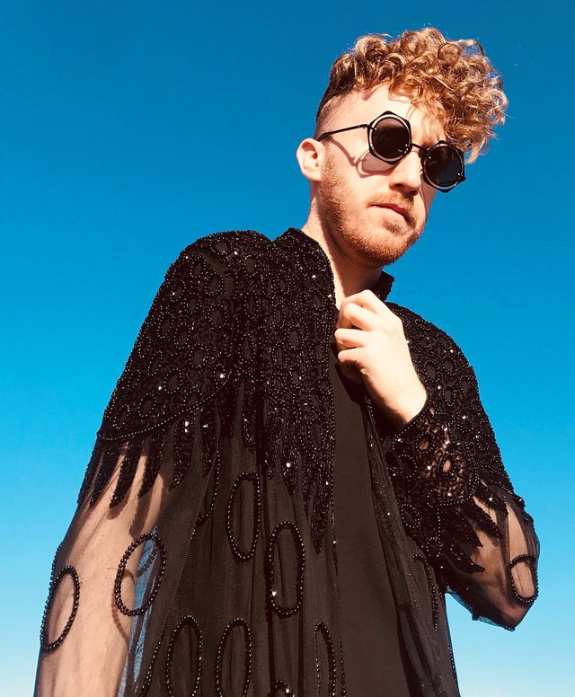 Daley in dark sunglasses and a beaded black cape against a clear blue sky