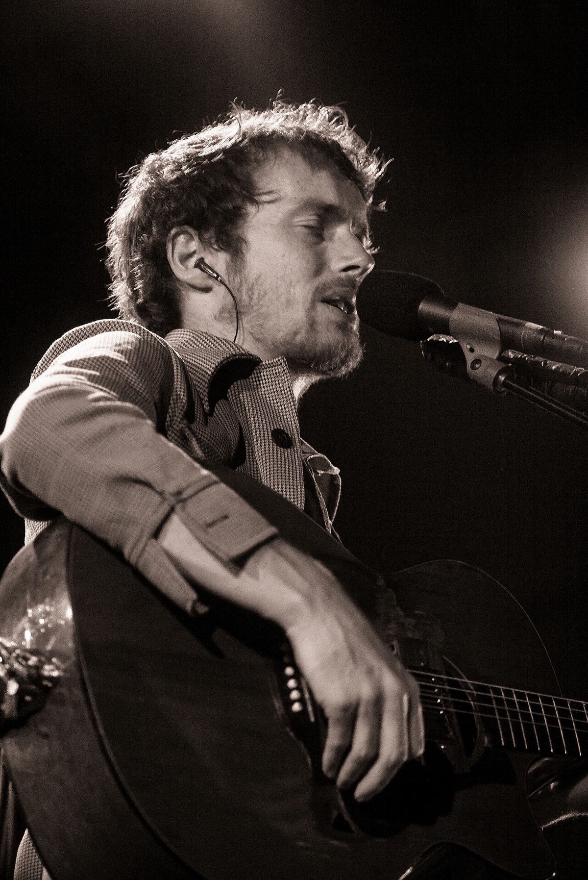 Black and white photo of Damien Rice singing into a microphone while playing an acoustic guitar