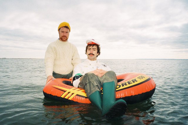 Dana and Alden in wool sweaters and rain boots, one sitting in an orange inflatable raft on open water