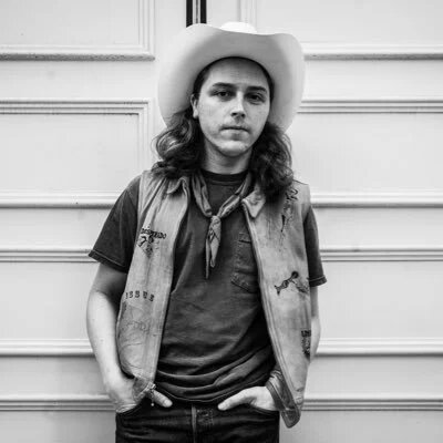 Black and white portrait of a long-haired man in a cowboy hat, scarf and vest leaning against a paneled wall