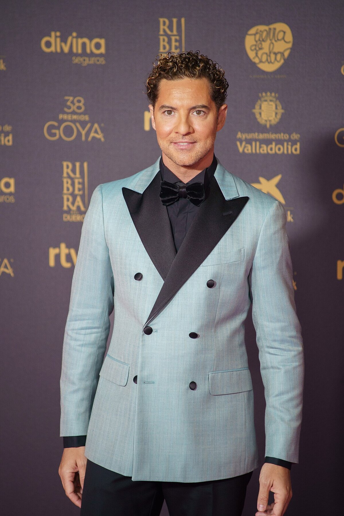 David Bisbal in a light blue double-breasted jacket and black bow tie, posing in front of a sponsor backdrop