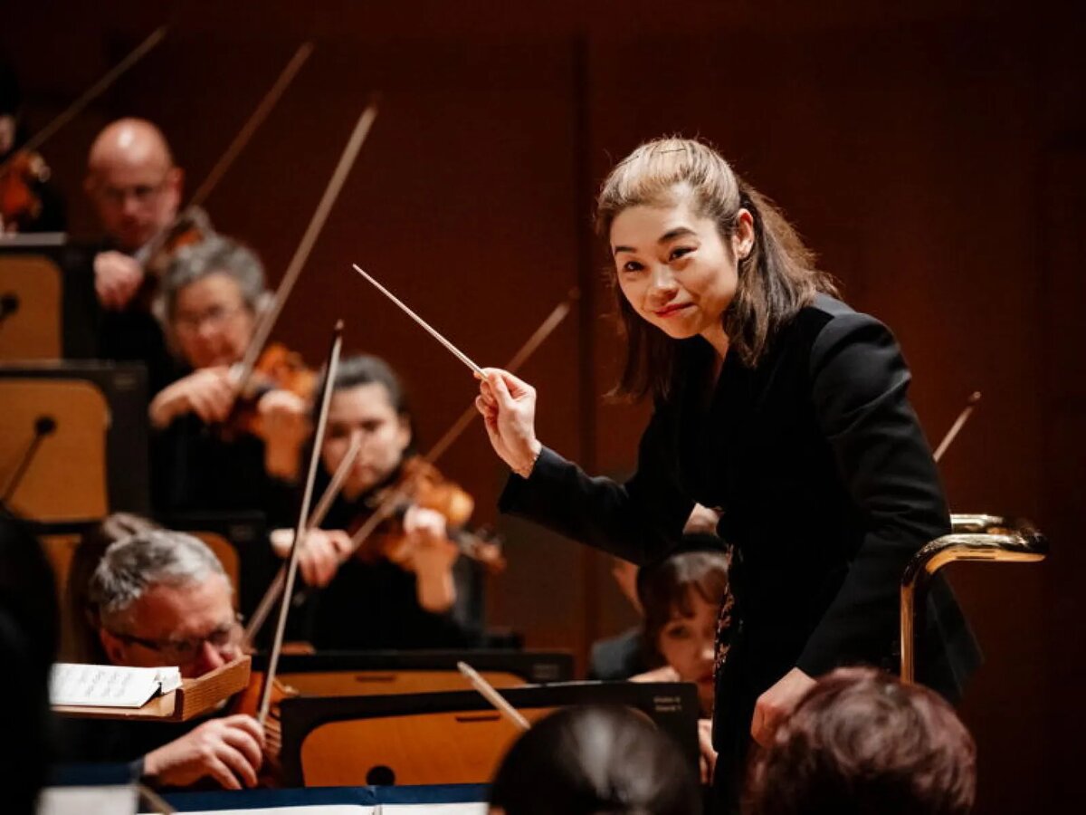 Conductor Elim Chan smiling and leading orchestra musicians, baton raised