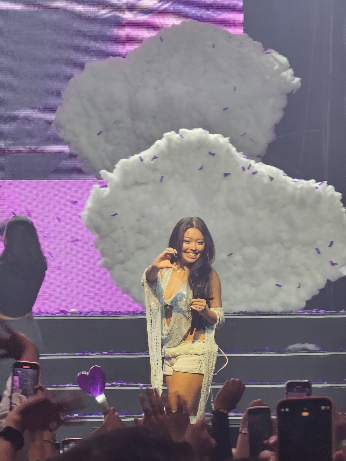 Denise Julia smiling on stage and making a heart gesture in front of a large cloud-shaped set piece