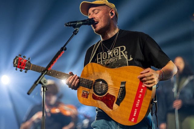 Dermot Kennedy singing into a microphone while playing an acoustic guitar under blue stage lights