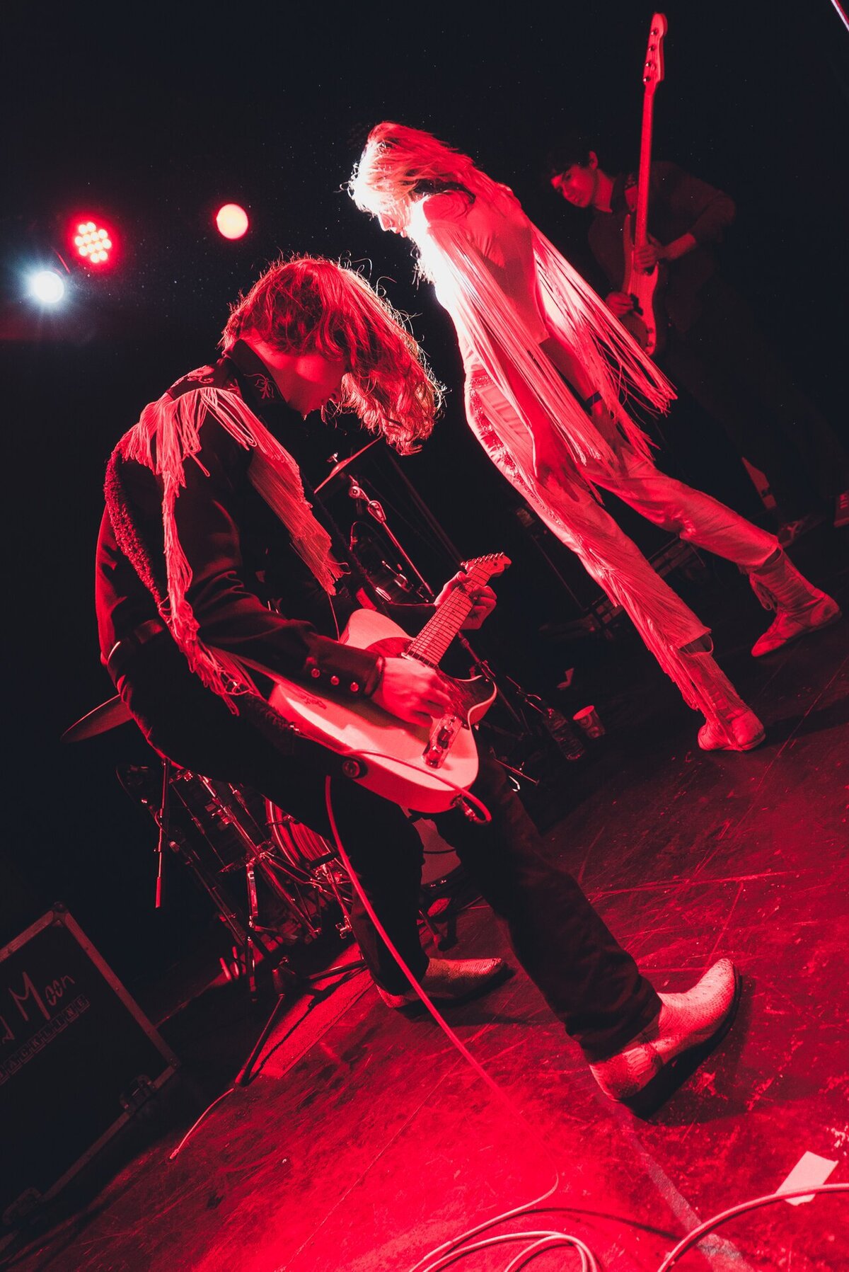 Two members of a rock band under red stage lights, one hunched over a guitar and one in a fringed outfit